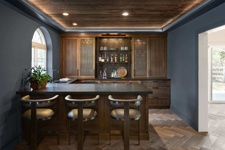 Dark blue home bar with wood cabinetry, herringbone floors, and a backlit tray ceiling.